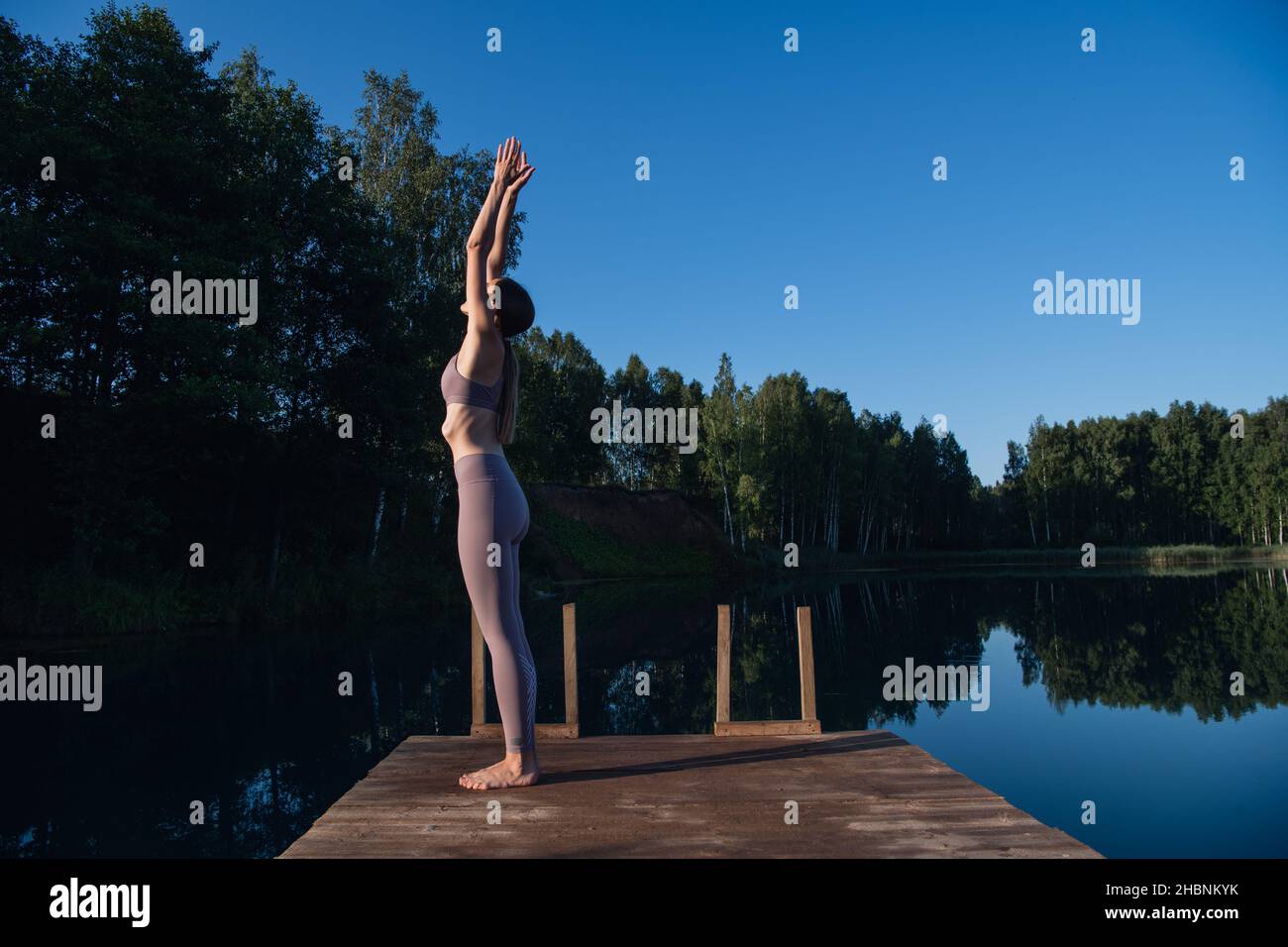 Young female practicing yoga against the sky on a sunrise. Pretty slim woman performs an exercise. Healthcare, meditaion, welness, mindfulness concept Stock Photo