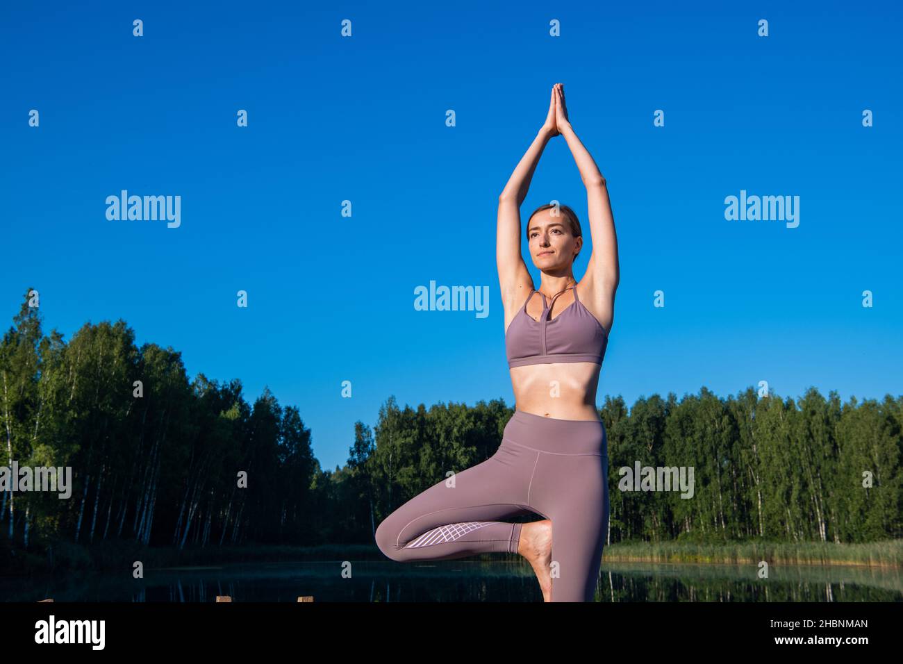 Young female practicing yoga against the sky on a sunrise. Pretty slim woman performs an exercise. Healthcare, meditaion, welness, mindfulness concept Stock Photo