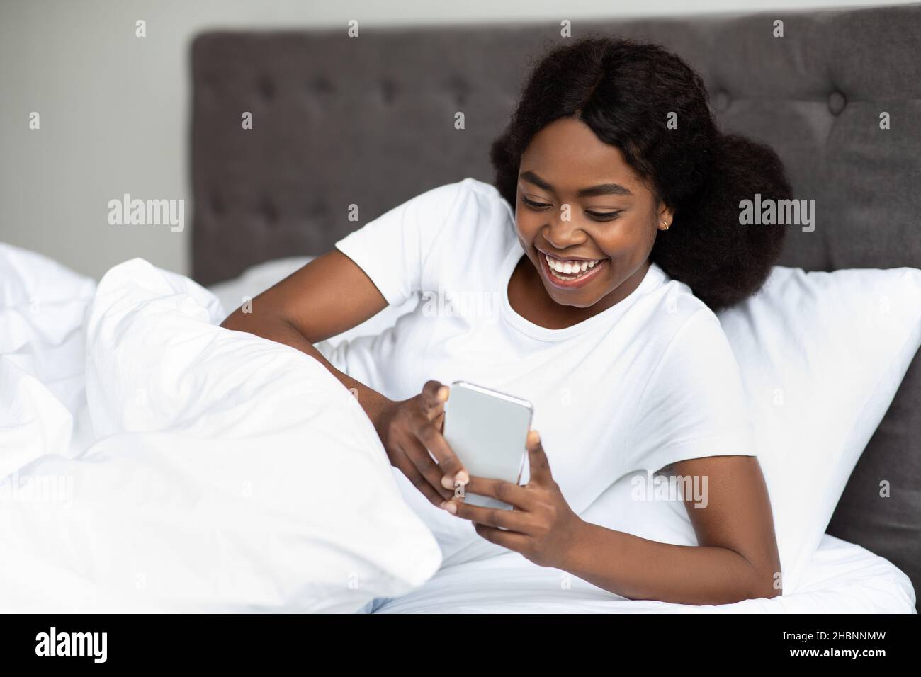 Smiling black lady using cellphone while laying in bed Stock Photo