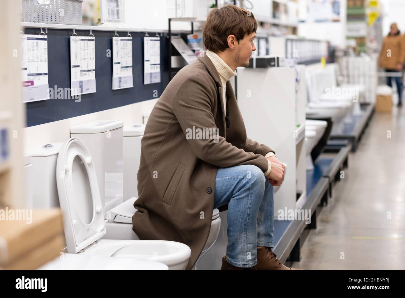 European man in a brown coat is sitting on the toilet in a hardware store, choosing a toilet for the bathroom during repairs Stock Photo