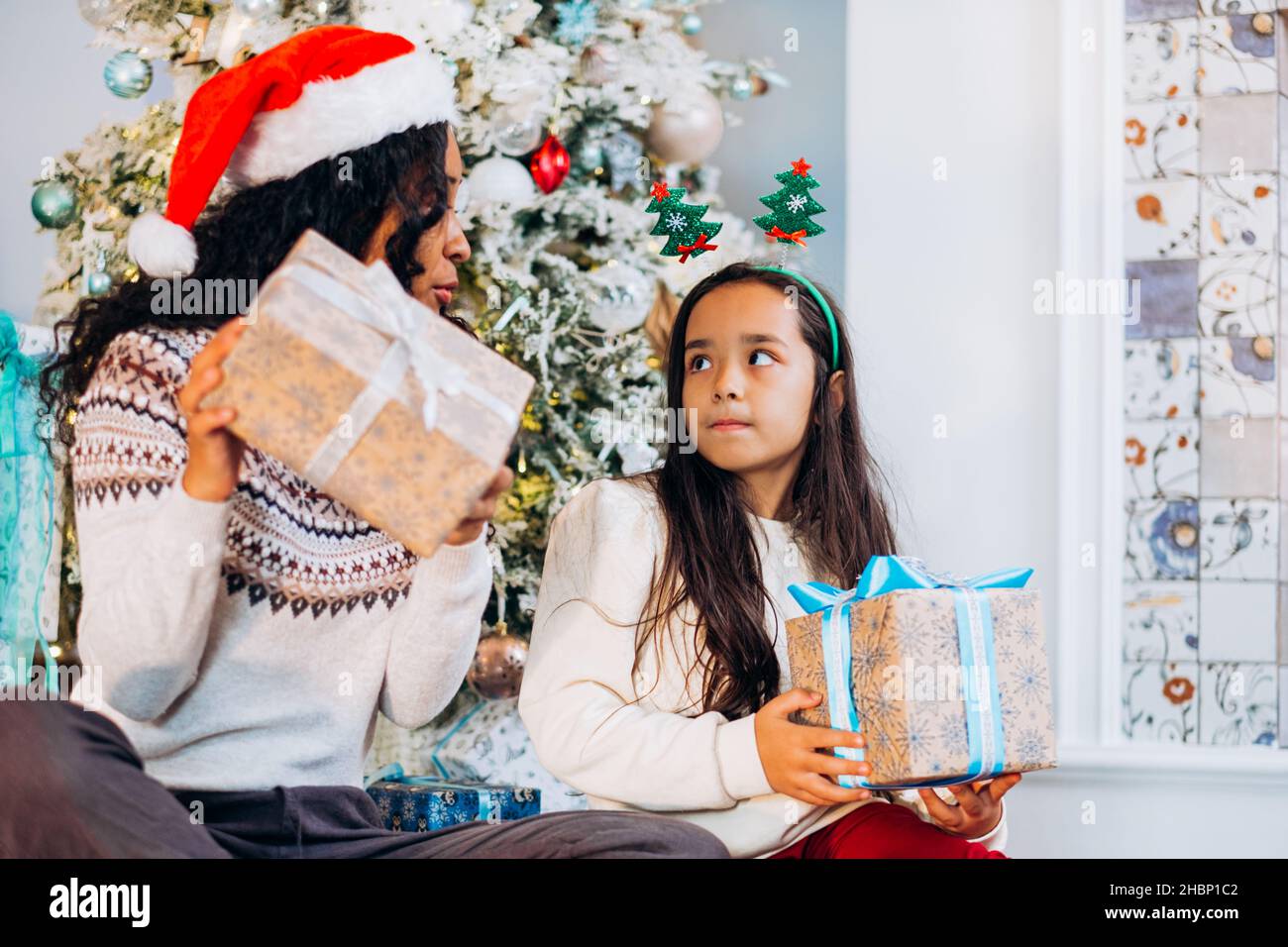 Curly long-haired African American mother and brunette daughter sit on carpet and open festive presents smiling happily near Christmas tree Stock Photo