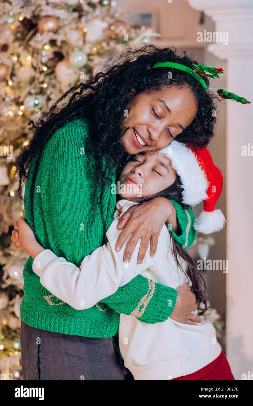African American woman with curly hair and brunette daughter pose hugging and wearing Christmas hats against tree decorations Stock Photo