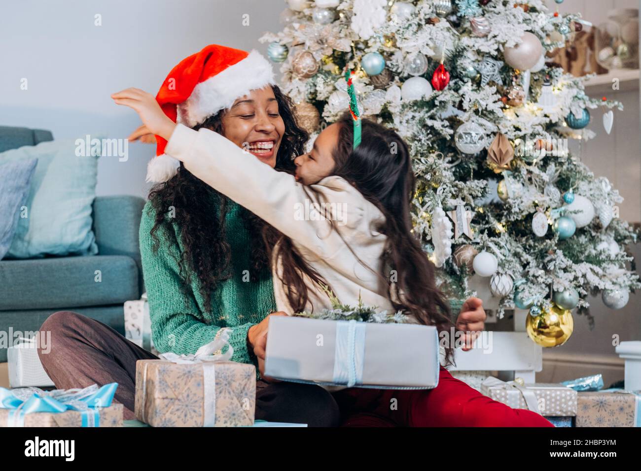 African American mother and brunette long-haired daughter in festive hats hug tightly smiling sitting among gift boxes by Christmas tree Stock Photo