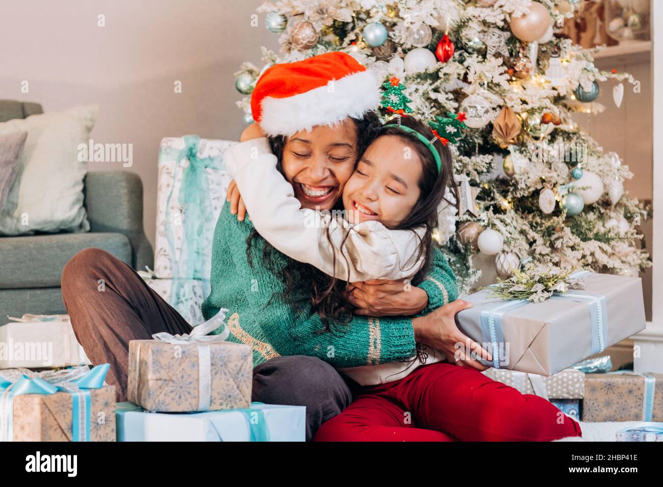 African American mother and brunette long-haired daughter in festive hats hug tightly smiling sitting among gift boxes by Christmas tree Stock Photo