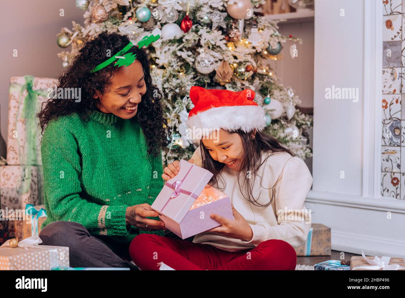 Curly African American woman and brunette long-haired daughter wearing holiday hats open present box sitting against Christmas tree Stock Photo