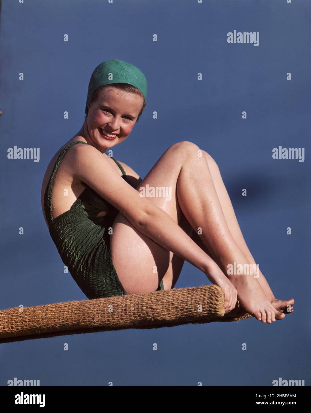 Young woman in green, 1-piece bathing suit with green bathing cap sitting on a diving board with knees pulled up Stock Photo