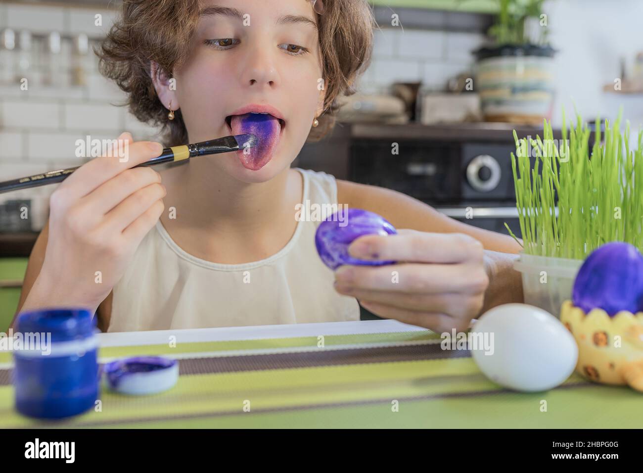 Child girl paints her tongue with food paints. child teenage girl holds an art brush and an Easter egg in her hand. DIY Easter eggs paint. Stock Photo