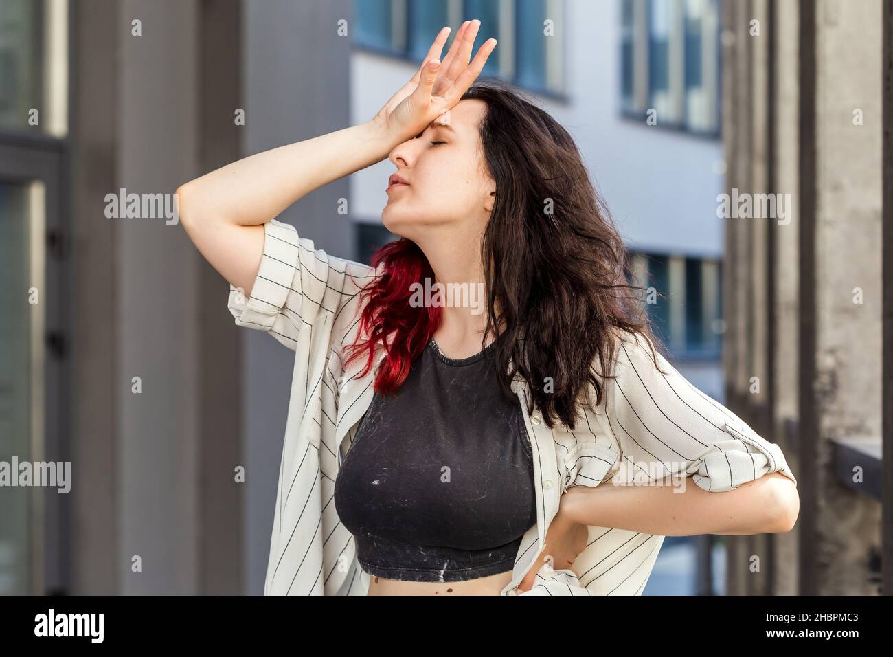 Waist-up shot of white european woman, who is holding arm on forehead showing despair, over office building background. Negative emotion concept. Pers Stock Photo