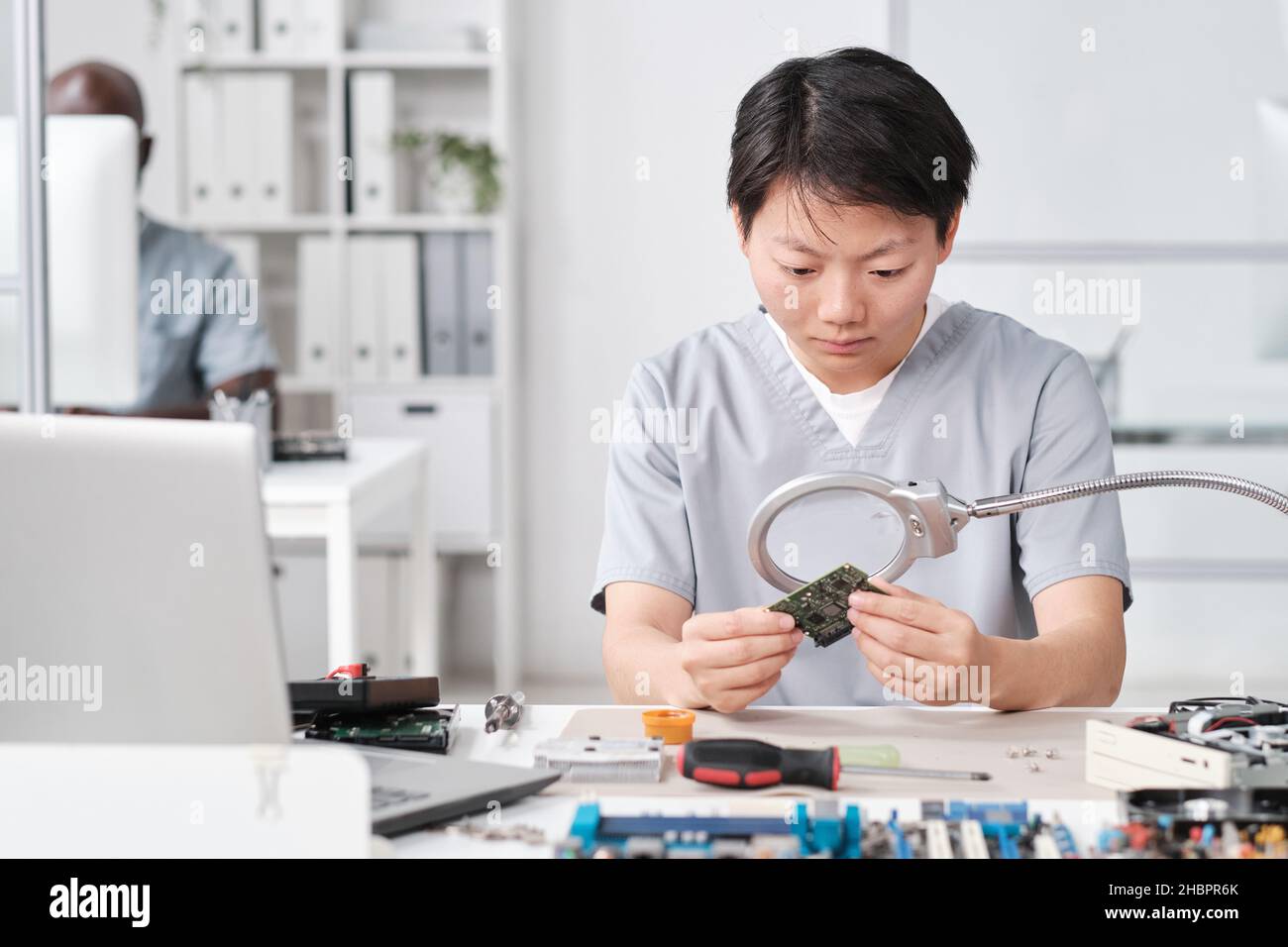 Young Chinese female in uniform looking at microchip in board through magnifying glass while sitting by desk Stock Photo