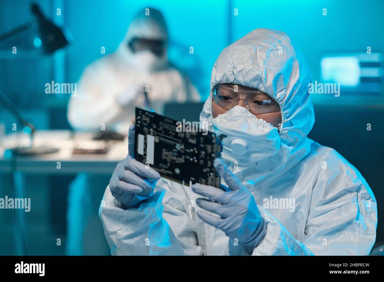 Young Asian female in protective workwear looking at circuit board in her gloved hands while working in laboratory Stock Photo
