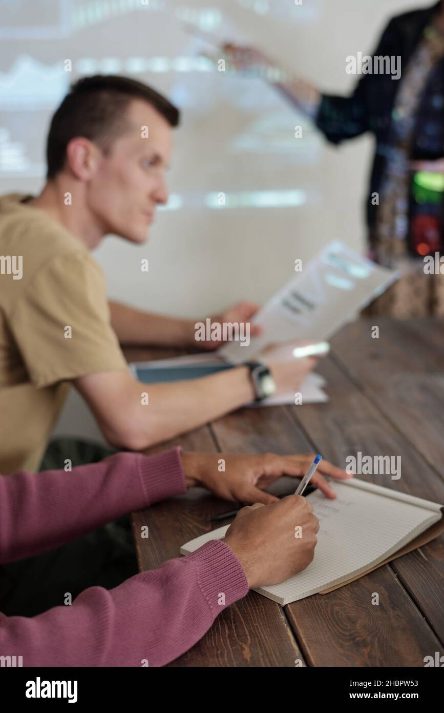 Hands of young biracial businessman making notes in notepad while sitting by table during presentation at seminar or business training Stock Photo