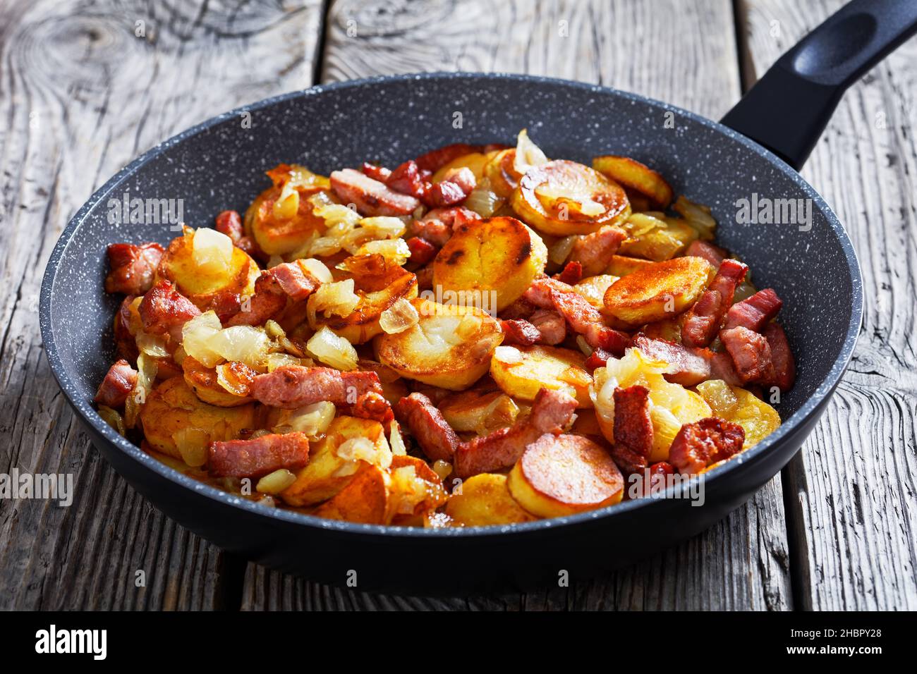 Bratkartoffeln, German fried Potatoes with Bacon and onion in a skillet on a wooden rustic table, close-up Stock Photo