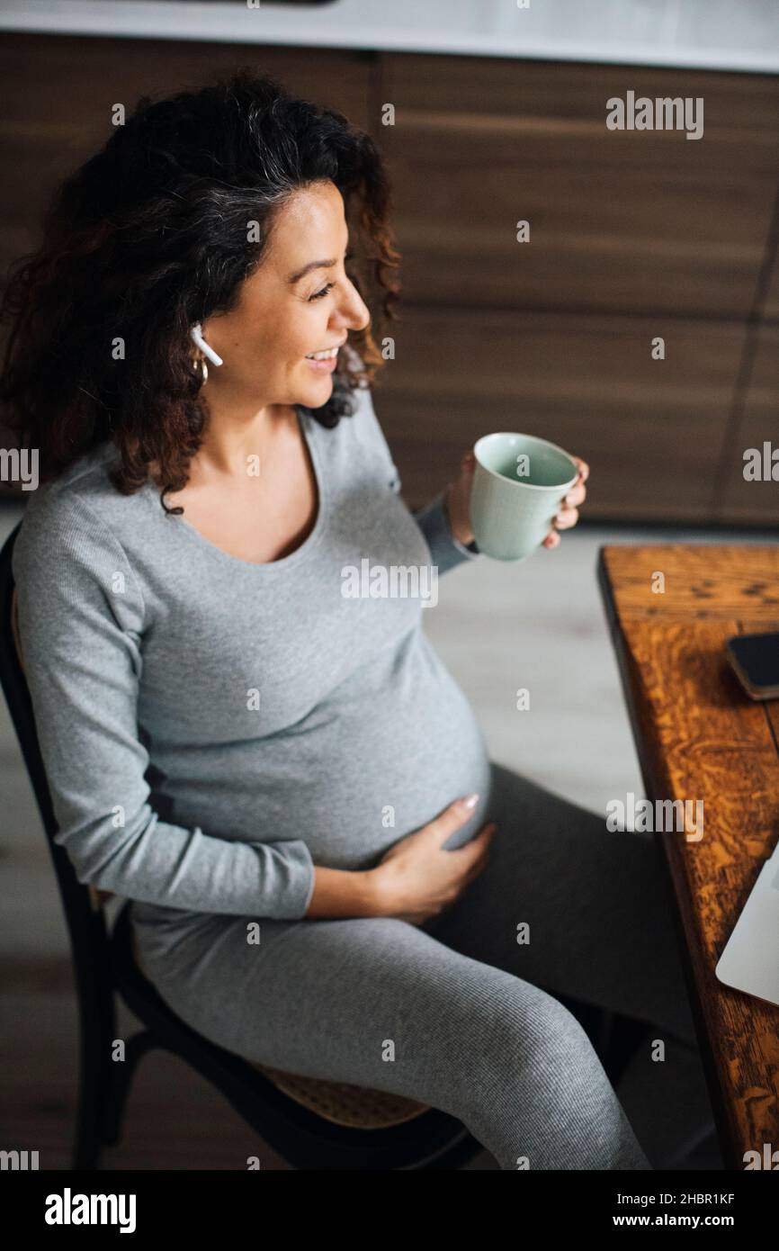 Pregnant female freelancer talking through bluetooth while having coffee at home Stock Photo