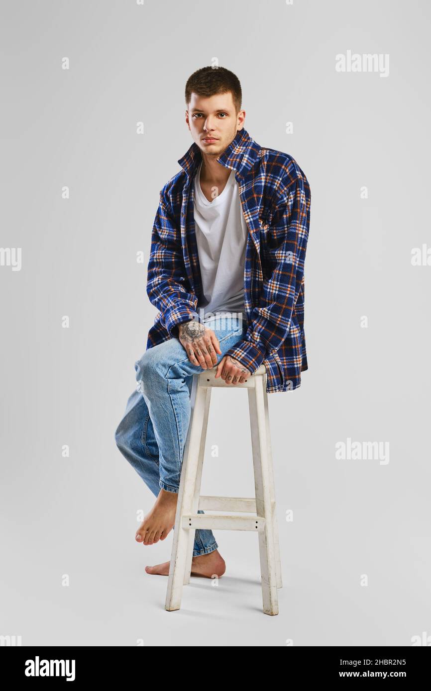 Full length studio portrait of young cocky man in shirt and jeans sitting on tall wooden chair Stock Photo