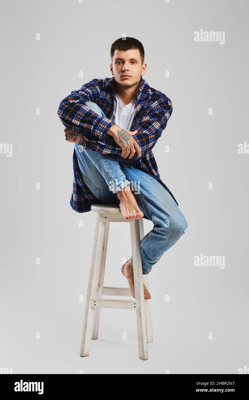 Full length studio portrait of young barefoot man in shirt and jeans sitting on tall wooden chair Stock Photo