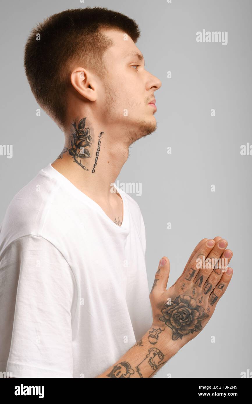 Young tattooed manstanding in profile, holds palms of his hands together and praying Stock Photo