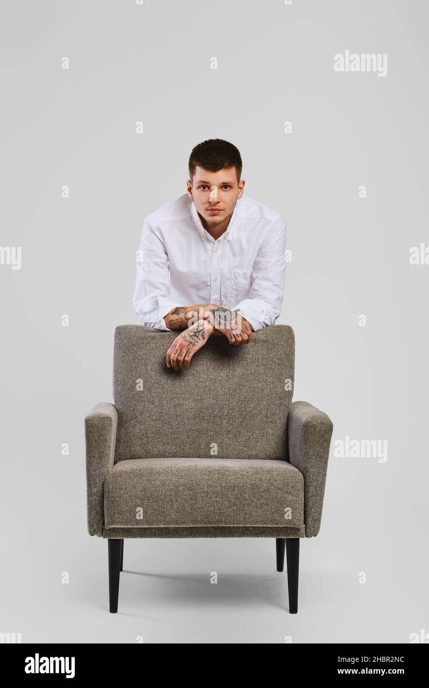 Full length studio portrait of young barefoot man in white shirt and ivory trousers leaning to armchair Stock Photo