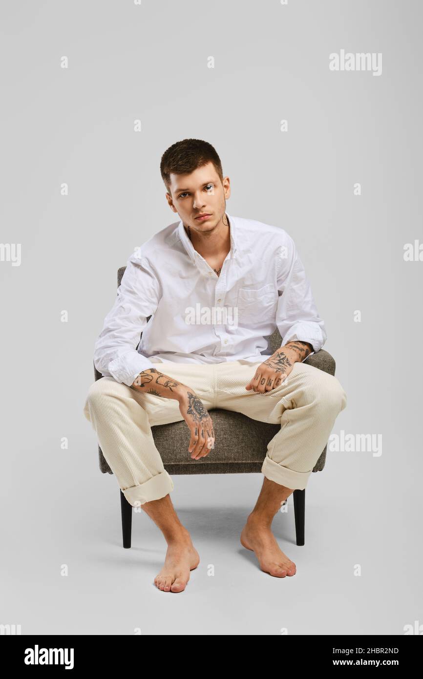 Full length studio portrait of young barefoot man in white shirt and ivory trousers sitting in armchair Stock Photo