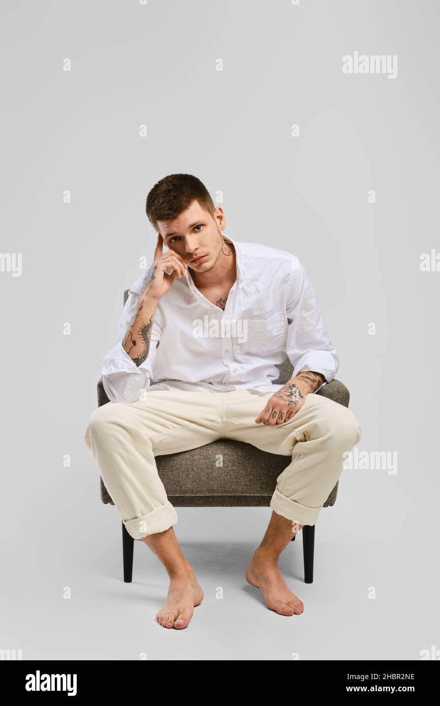 Full length studio portrait of young barefoot man in white shirt and ivory trousers sitting in armchair Stock Photo