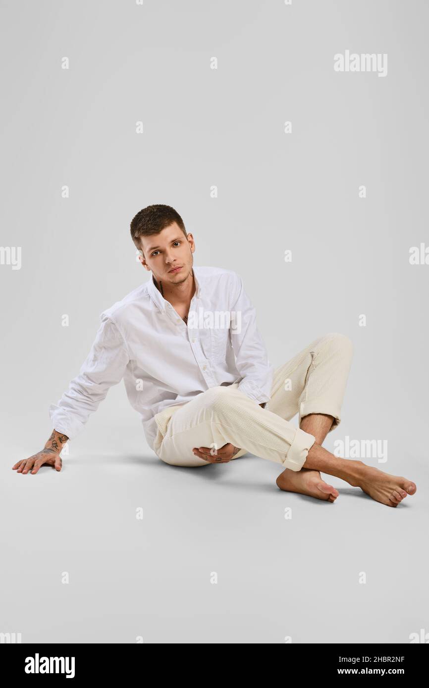 Full length studio portrait of young barefoot man in white shirt and ivory trousers sitting the floor Stock Photo