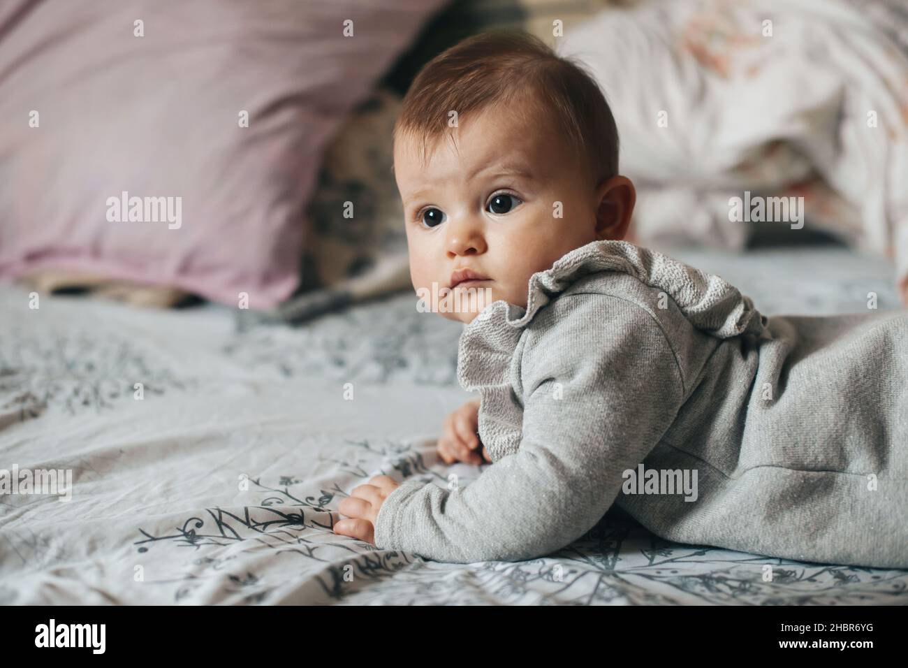 Side viewof a little girl lies on her stomach on the bed and looking at the camera. New born child. Beautiful girl. Healthy lifestyle. Stock Photo