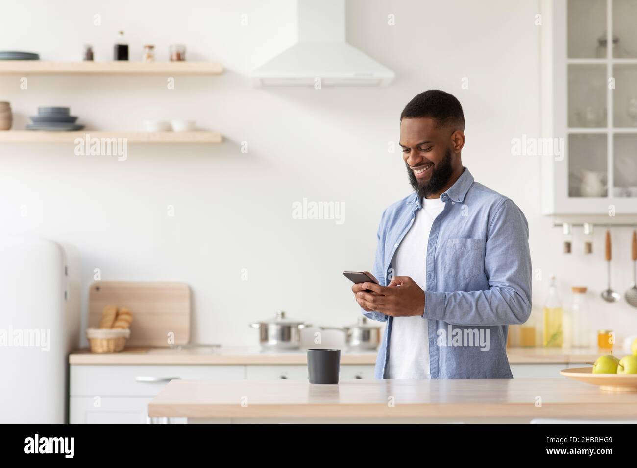 Smiling young bearded african american guy in minimalist kitchen interior reads news Stock Photo