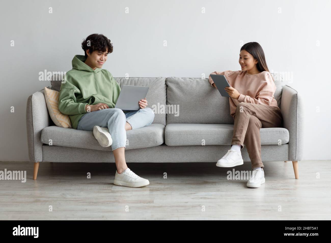 Handsome young Asian guy and his girlfriend using modern gadgets while sitting on couch at home, copy space Stock Photo