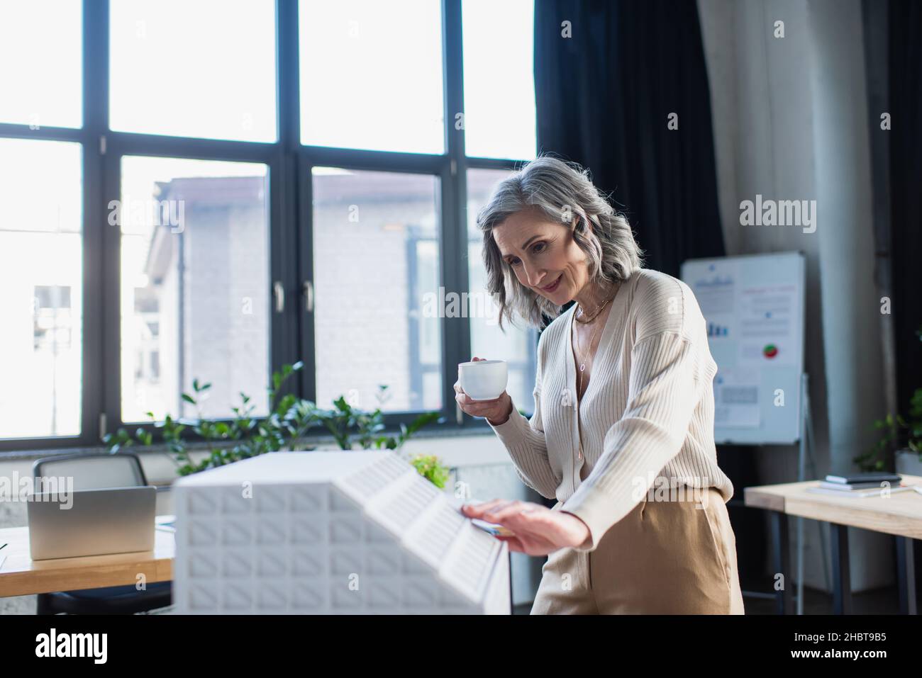 Cheerful mature businesswoman holding cup near model of building in office Stock Photo