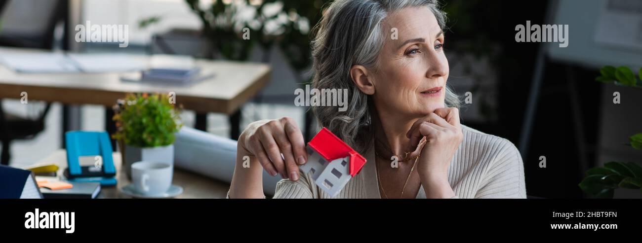Pensive businesswoman holding model of house in office, banner Stock Photo