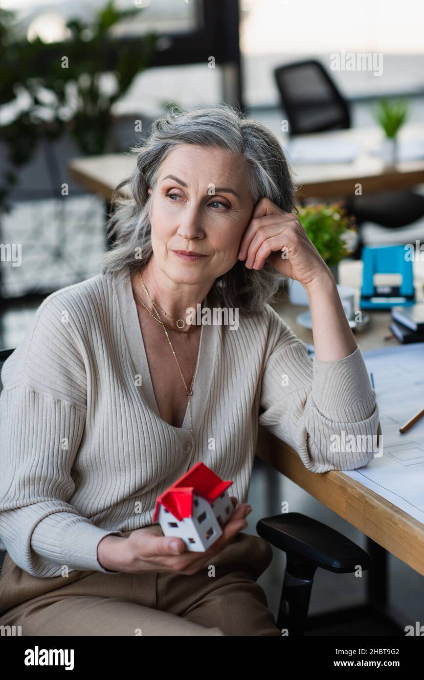 Thoughtful businesswoman holding model of house near blueprint on table in office Stock Photo