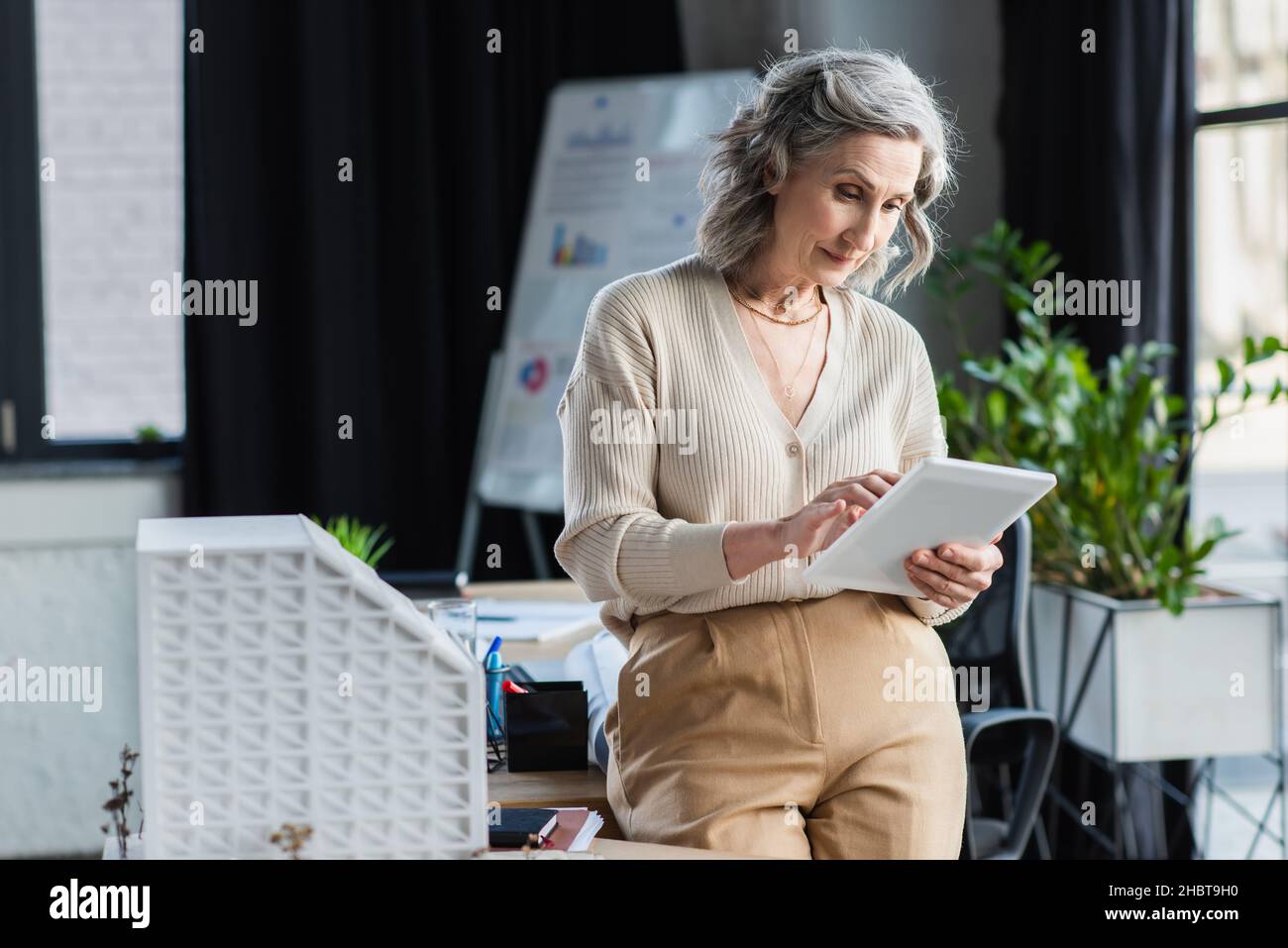 Grey haired businesswoman using digital tablet near model of building in office Stock Photo