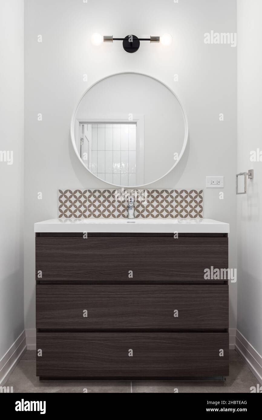 A wooden bathroom vanity cabinet with marble countertop, tiled backsplash, and a light over a circular mirror. Stock Photo