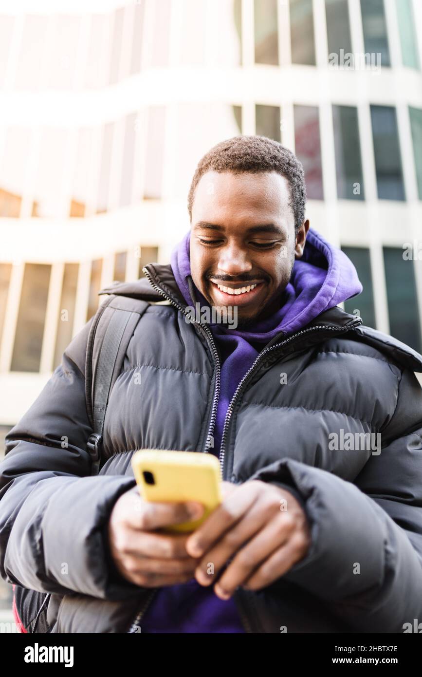 Positive black male in outerwear smiling and reading messages on smartphone while standing against contemporary skyscraper in city Stock Photo