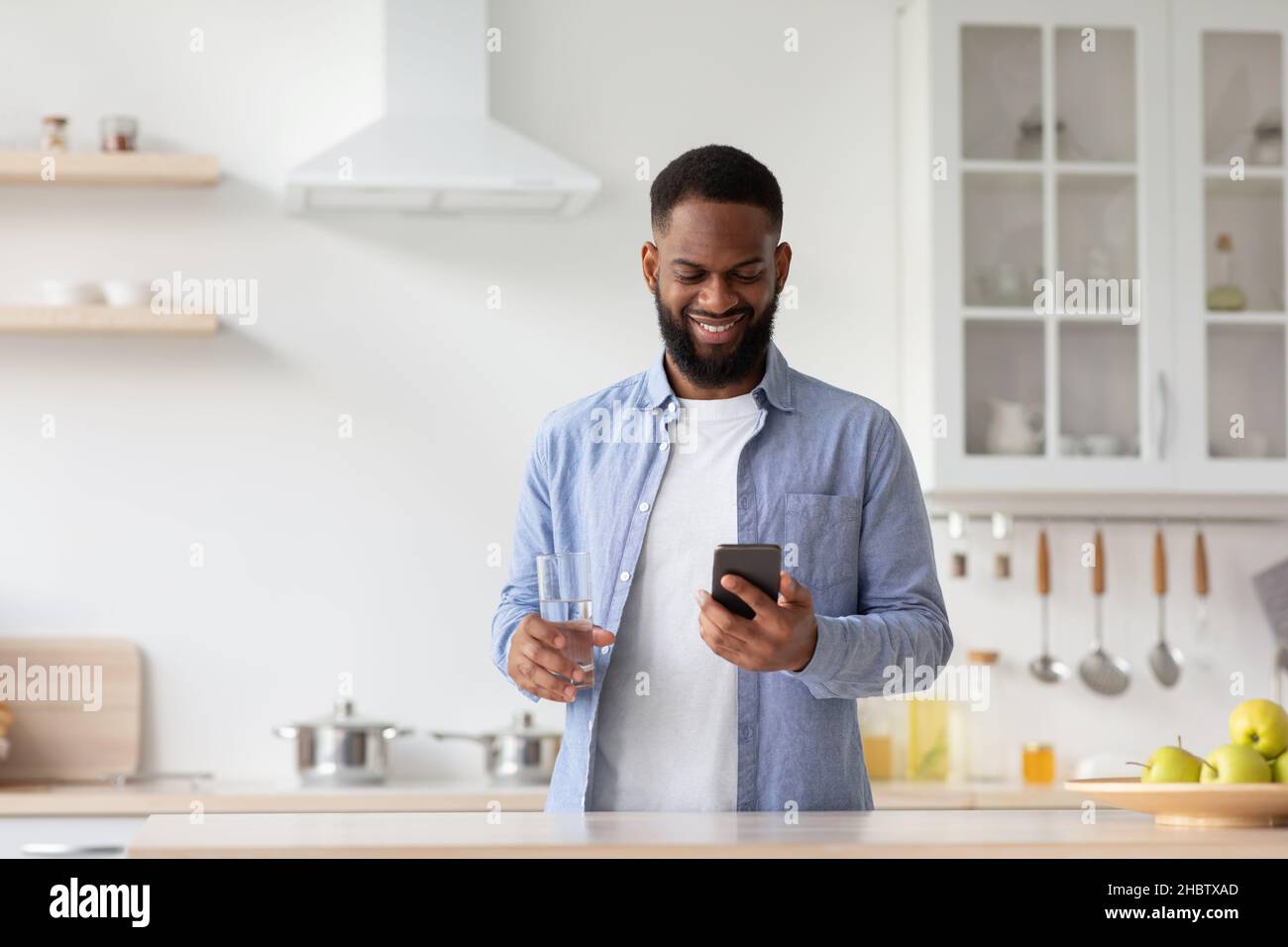 Happy young black man with glass of clear water in hand in minimalist kitchen interior, chatting on phone Stock Photo