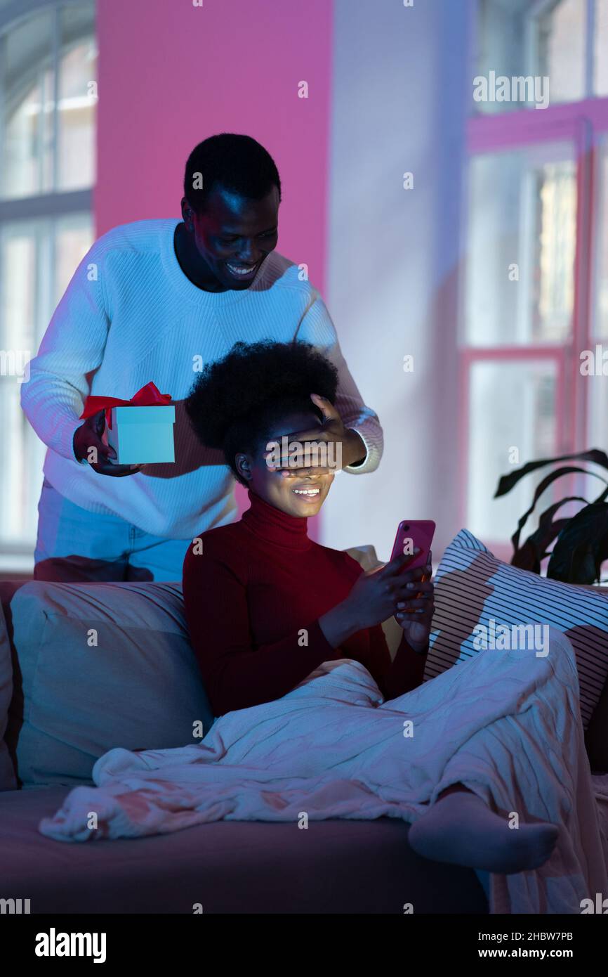 Afro man surprise girlfriend with gift. Woman sit on couch, closed eyes receive unexpected present Stock Photo