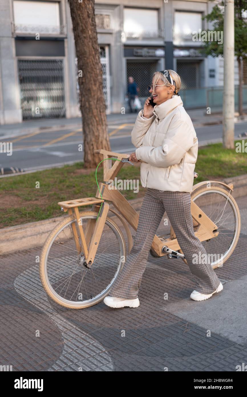 Full body cheerful adult woman in outerwear smiling and answering phone call while walking near wooden eco bike on pavement in city. Stock Photo