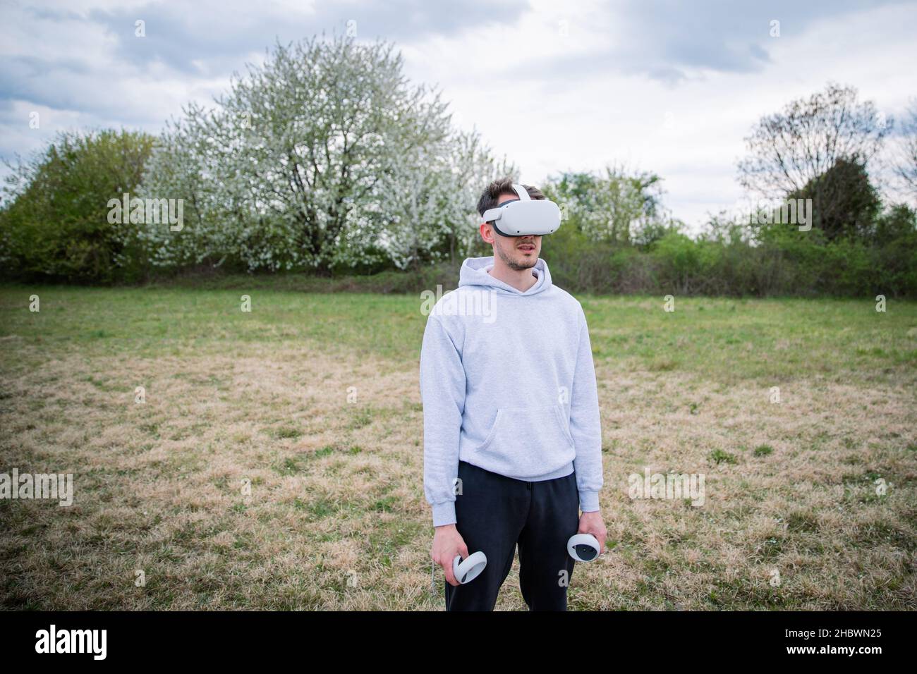 A boy uses VR headsets to play in the metaverse, he holds two controllers and he is in an open-air park. Stock Photo