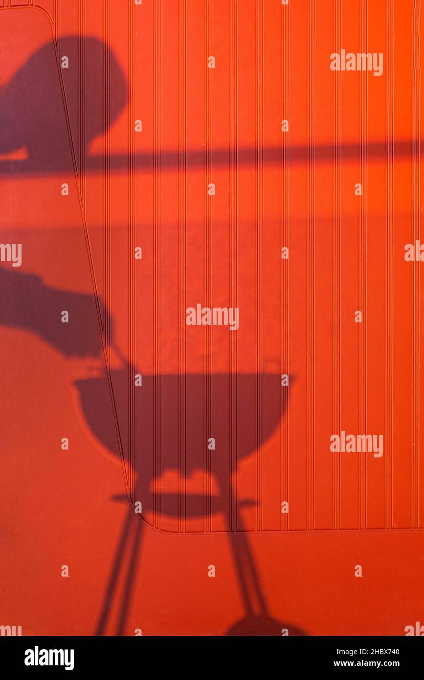 Shadow of person having a smoking barbecue against red wall. Shadow of head and hands of a man grilling meat on a smoking, hot bowl bbq on tripod Stock Photo