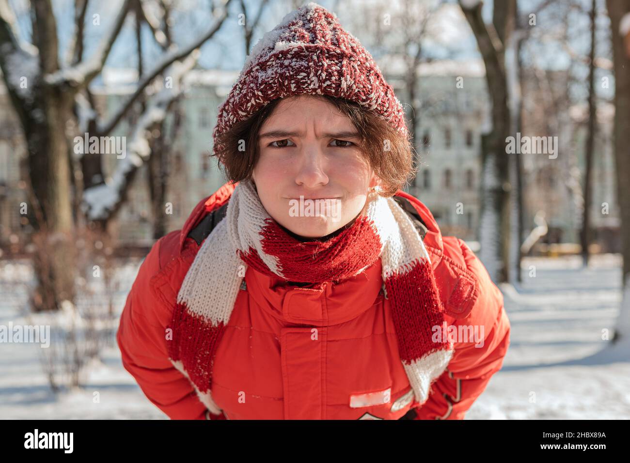 funny teenage girl in warm down jacket and knitted hat and scarf with red frostbitten cheeks and nose frowning with displeasure looks into camera Stock Photo