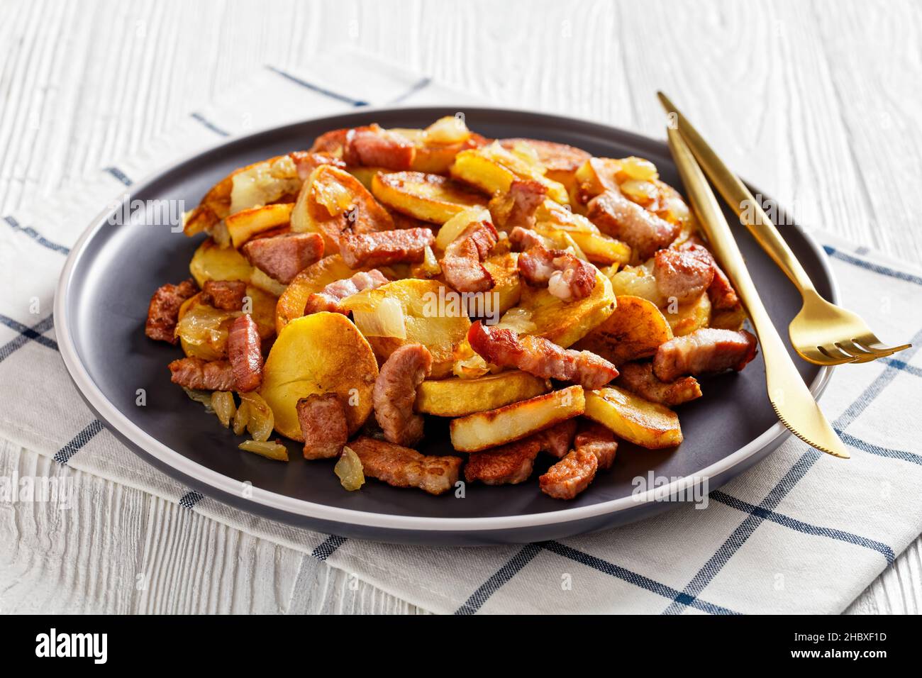 Bratkartoffeln, fried Potatoes with Bacon and onion on a plate with cutlery on a white wooden table, German cuisine Stock Photo