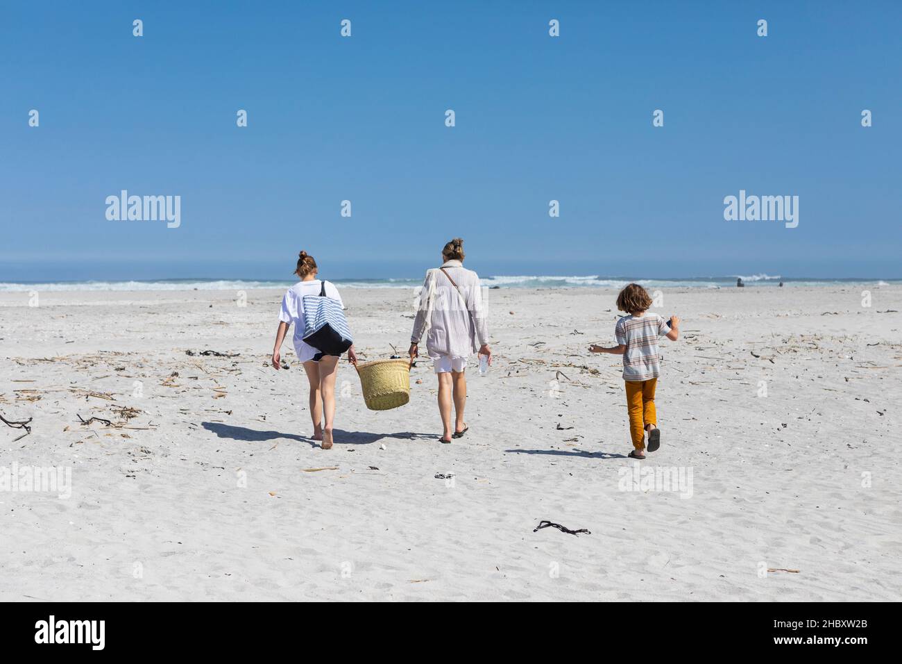 Mother and teenage daughter walking on a sandy beach carrying a basket, boy following. Stock Photo