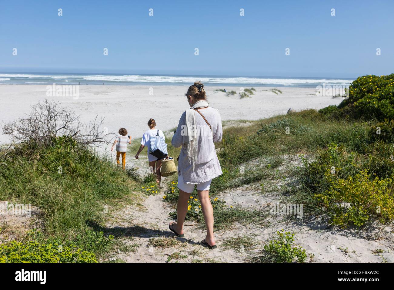 A family walking through the sand dunes towards the ocean with baskets and bags. Stock Photo