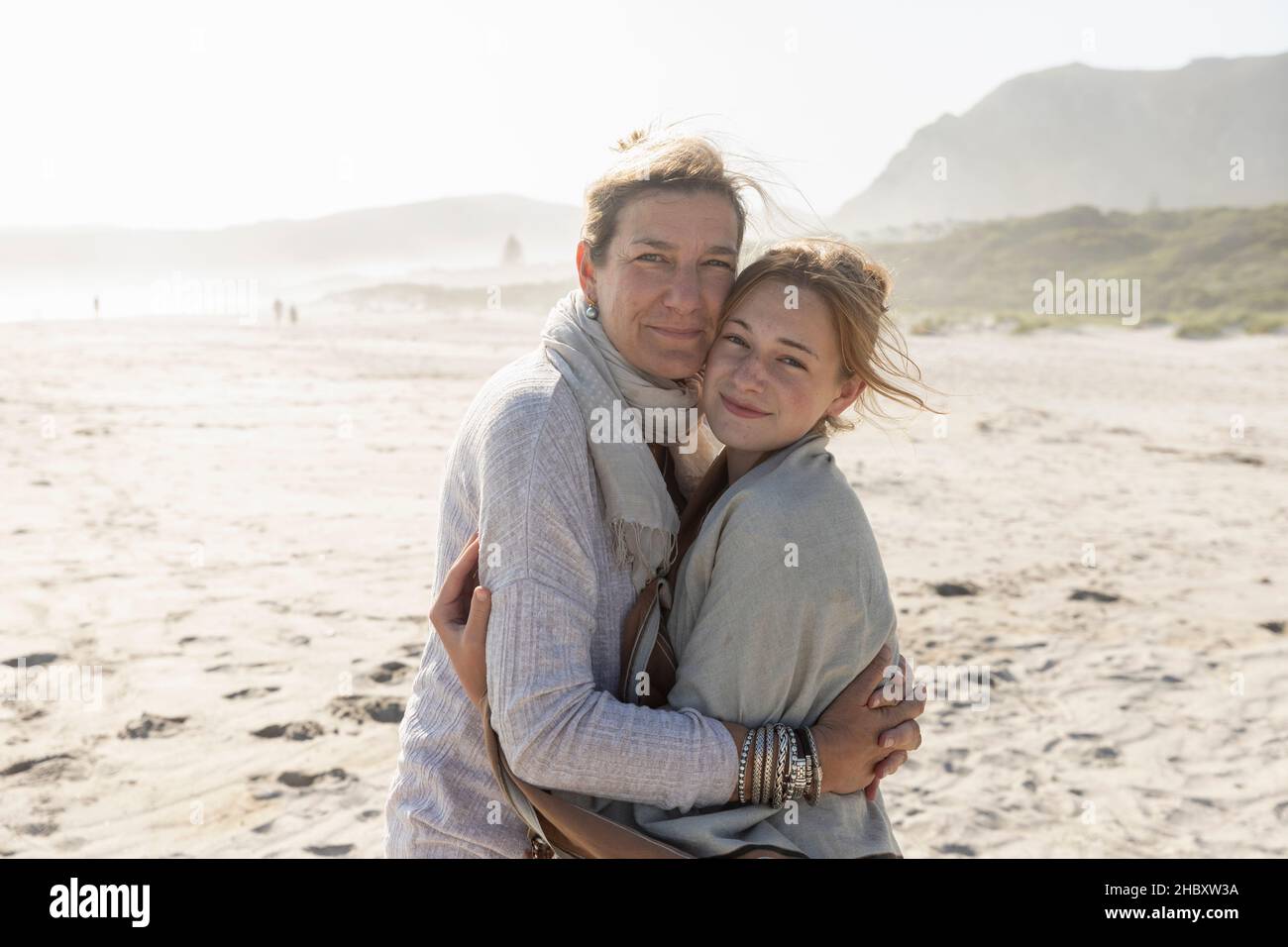 Adult woman and her teenage daughter hugging, standing on a windswept beach Stock Photo