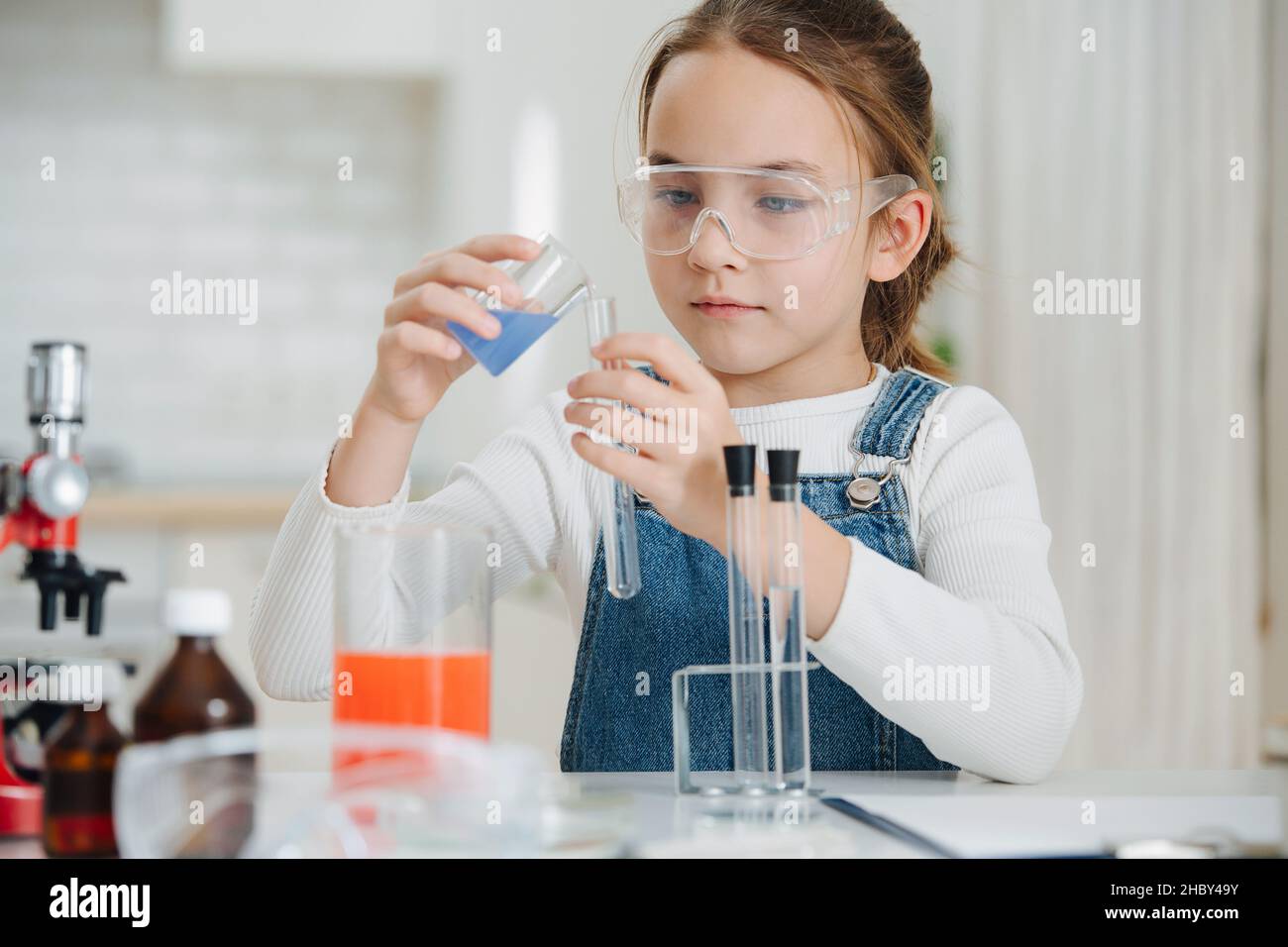 Cute focused girl doing home science project, pouring liquid into a flask. She is wearing plastic glasses and has glass chemical equipment and microsc Stock Photo
