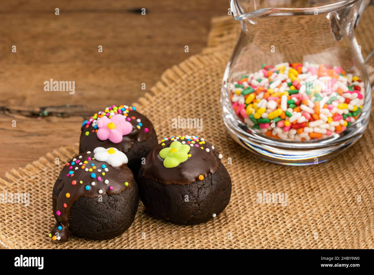 Chocolate balls topping with multicolored rainbow sprinkles and sugar flower on sack cloth and colorful rainbow sprinkles in transparent glass jug on Stock Photo