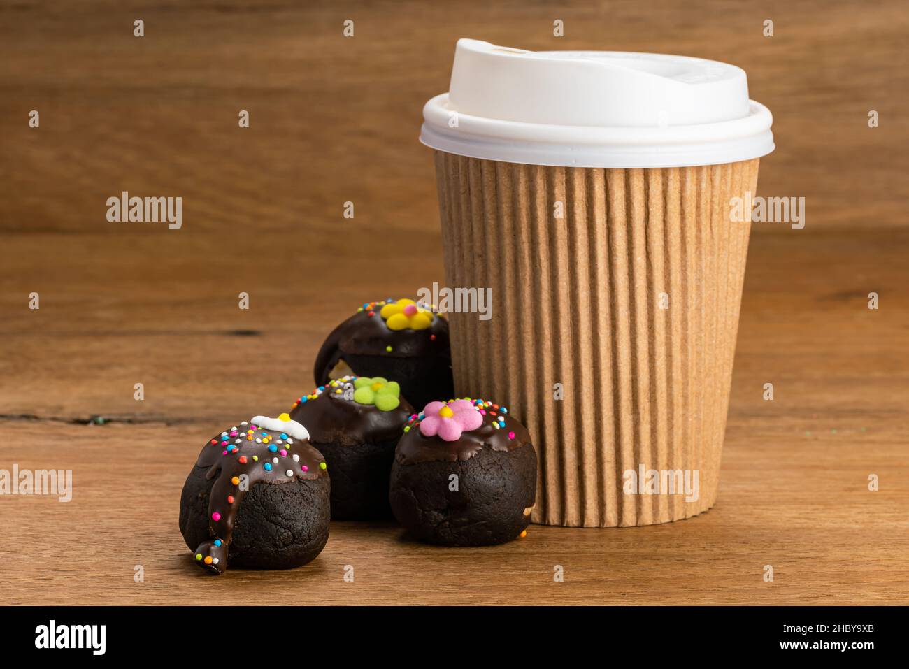 Chocolate balls or choc balls topping with multicolored rainbow sprinkles and colored sugar flower with paper cup of coffee on wooden table. Stock Photo