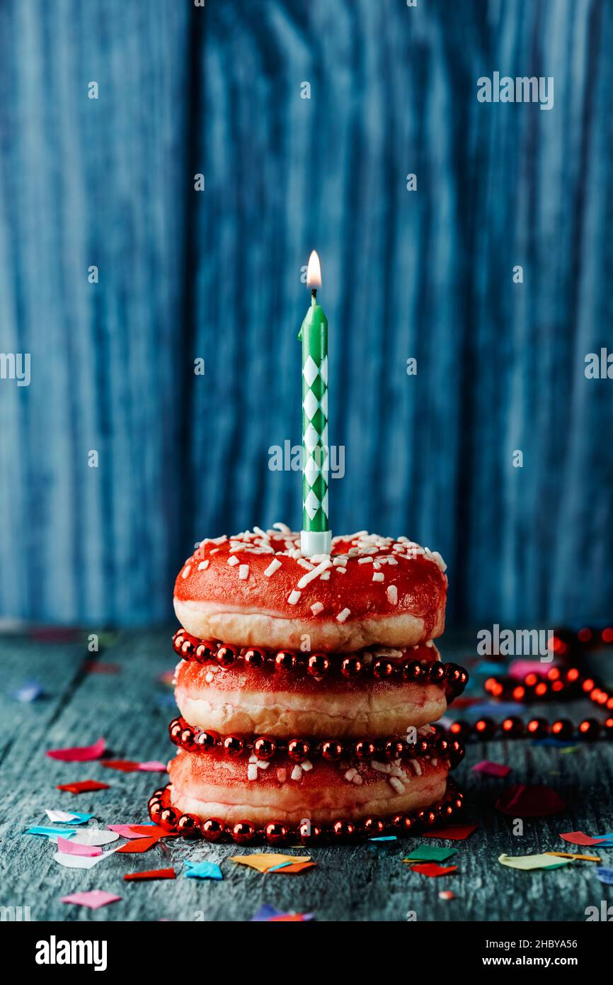 a pile of donuts, topped with a lit candle, on a gray rustic wooden table sprinkled with confetti of different colors Stock Photo