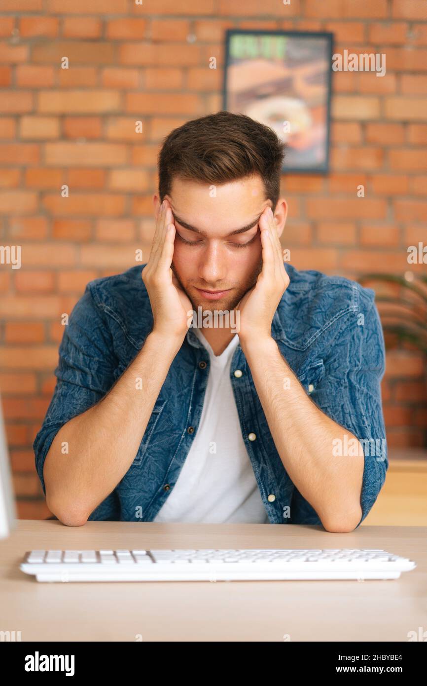 Vertical portrait of exhausted young man touching temples feeling unbearable headache sitting at desk with desktop computer at home office. Stock Photo