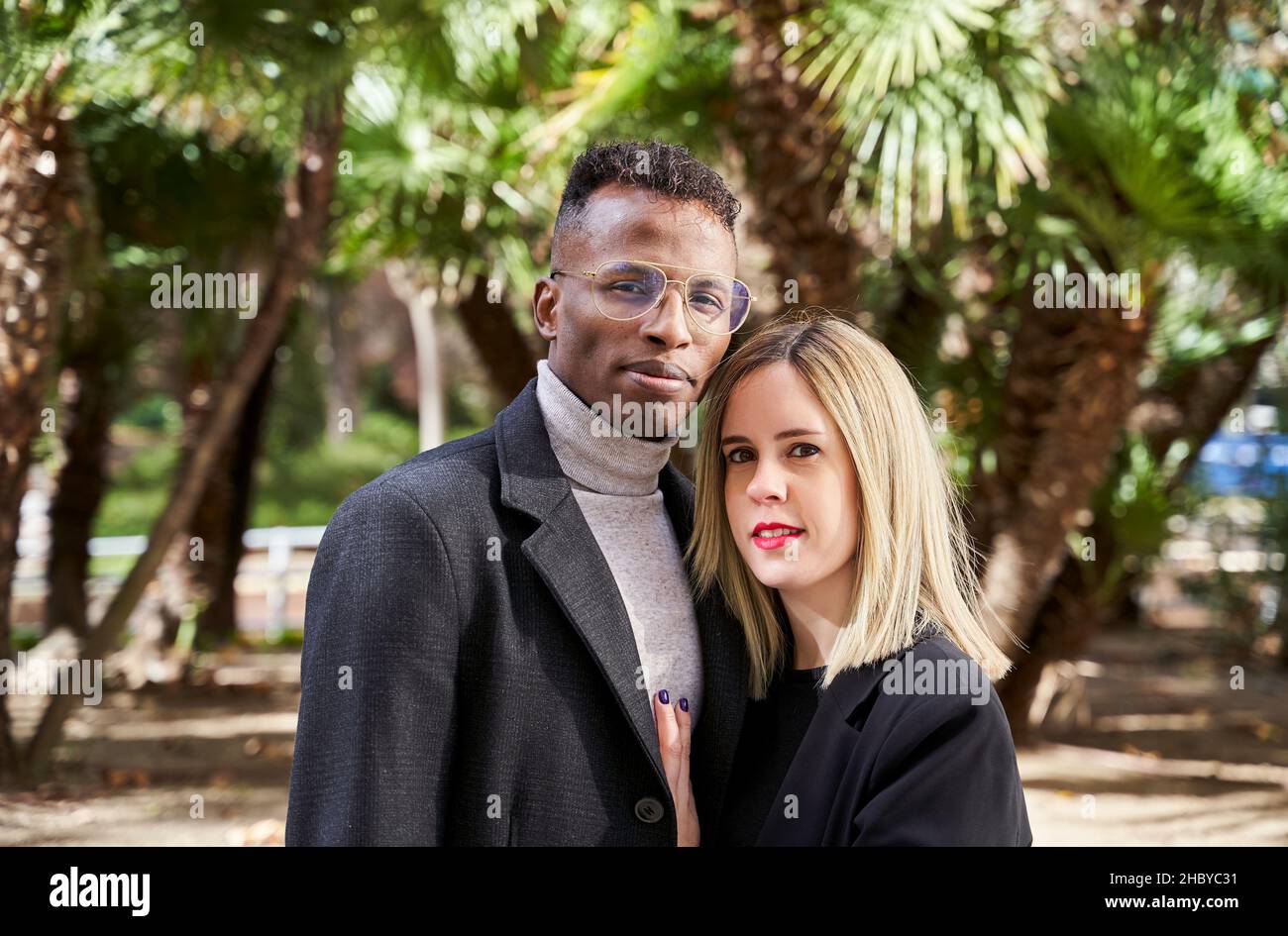 Black man and blond woman in outerwear looking at camera while standing against palms during romantic date on sunny day in exotic park Stock Photo