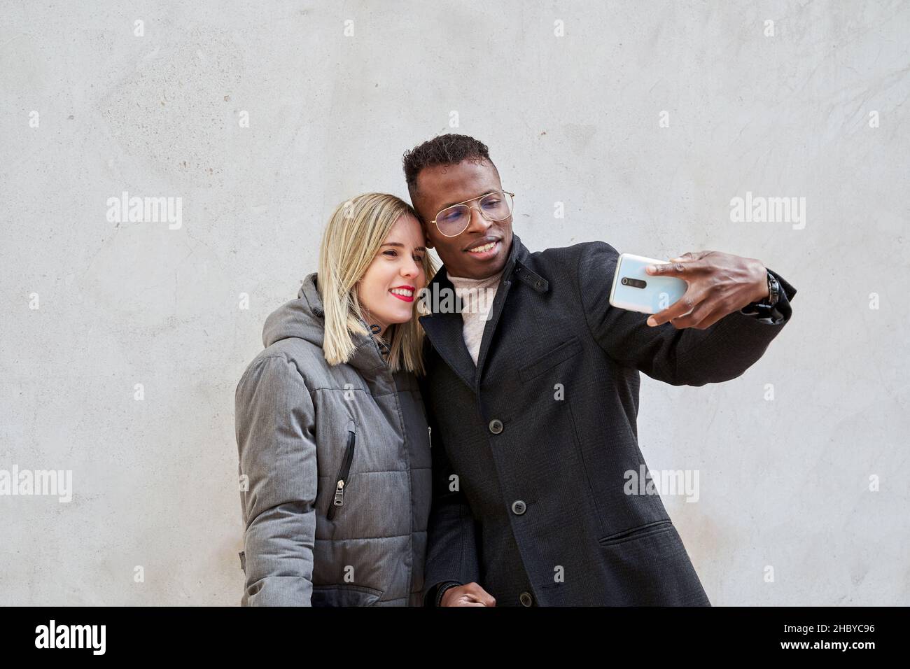 Diverse boyfriend and girlfriend in outerwear smiling and taking selfie against gray wall during date on city street Stock Photo